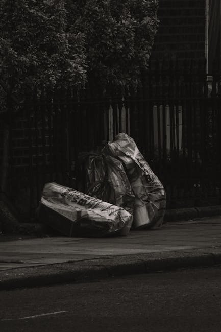 On a paved urban sidewalk, two large black plastic garbage bags are placed next to a wooden fence with vertical slats. The bags are tightly packed and crumpled, with visible creases and folds, suggest
